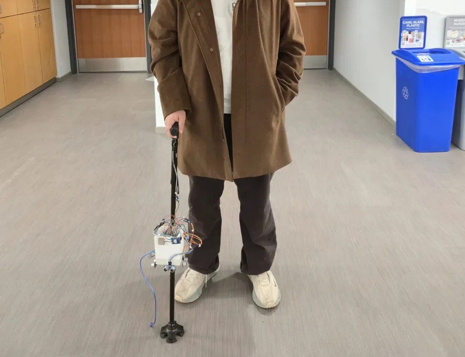 Rami holding the Smart Walking Stick during development; the electronics enclosure is open with the sensor wiring exposed, before final routing into the housing.